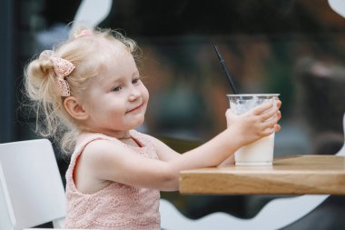 Sarışın ve milkshake, şehirdeki bir masada. Pembe elbiseli, at kuyruklu üç yaşında bir bebek kameraya duygusal pozlar veriyor..