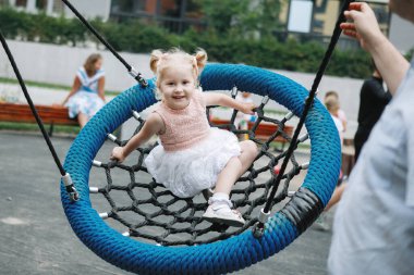 Salıncakta at kuyruklu küçük sarışın kız. Annem yardım eder ve gülümser..