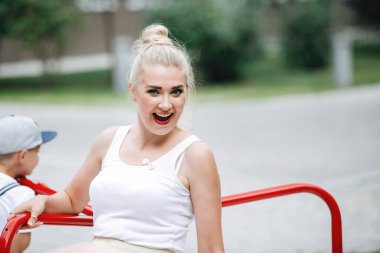 Sarışın bir annesi olan küçük bir çocuk oyun parkında atlıkarıncaya biniyor..