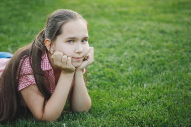 Genç kız çimlerin yeşil çimlerinde yatıyor. Dirseklerine yaslanmış, kameraya bakıyor. Pembe gömlek, uzun saç. Seçici odak, bulanık arkaplan.