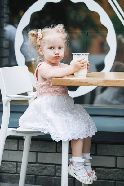 Sarışın ve milkshake, şehirdeki bir masada. Pembe elbiseli, at kuyruklu üç yaşında bir bebek kameraya duygusal pozlar veriyor..