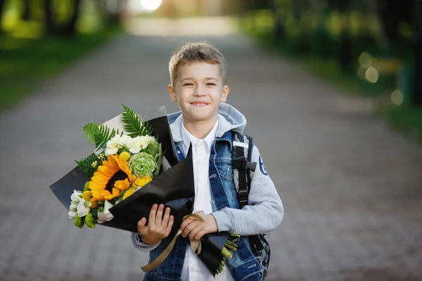 School teenager with a bouquet of flowers at the start of the school year in a white shirt and denim jacket with a hood in a city park. The first time in first class.