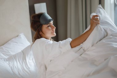 Beyaz cüppeli ve uyku maskeli bir kadın uyandı ve sabah bir otel odasında yatakta gülümsedi.