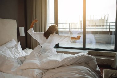 Beyaz cüppeli bir kadın pencerenin önündeki bir yatağa oturur ve sabahları bir otel odasında gerinir.
