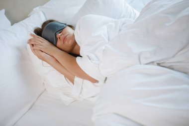 Yüzünde uyku maskesiyle beyaz bir yatakta uyuyan güzel bir genç kadın. Sağlıklı dinlenme.