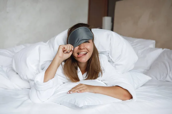 Woman in white bed with sleep mask, smiling and showing tongue. Rest at the hotel, travel tourists.