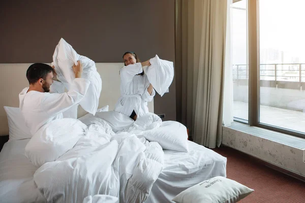 Woman and a man in a white bed are fighting a pillow in a bedroom with a huge window. Rest in the hotel, mating games.