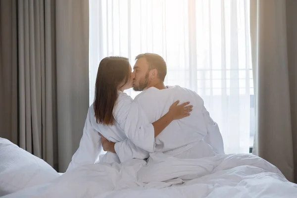 Lovers a man and a woman in white coats are sitting on the bed and kissing in front of the window in a hotel room. Back view.