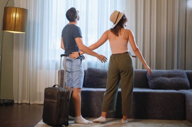 Şapkalı ve güneş gözlüklü bir adam ve kadın perdeli bir penceresi ve kanepesi olan bir otel odasına girdiler..