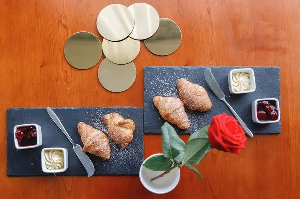 Serving breakfast on black square plates on the table. Red rose in a glass, croissants, jam and cutlery. Flat lay, top view.