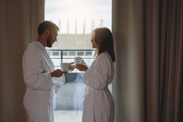 一个男人和一个女人穿着白色长袍 在卧室的窗户边喝咖啡 — 图库照片