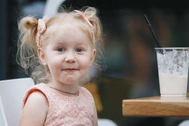 Sarışın ve milkshake, şehirdeki bir masada. Pembe elbiseli, at kuyruklu üç yaşında bir bebek kameraya duygusal pozlar veriyor..
