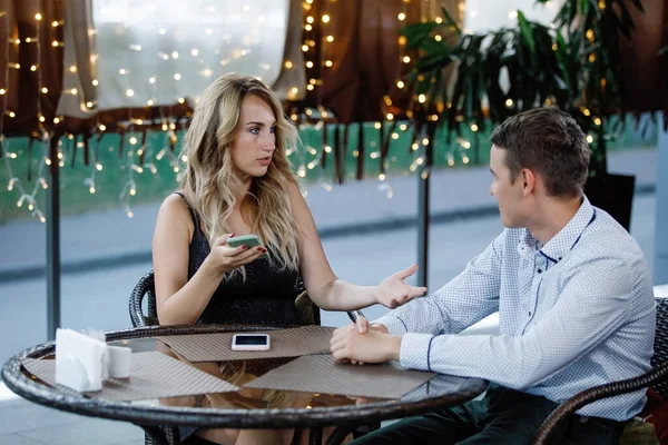 The couple\'s quarrel at the cafe table. The outraged woman shows the man a smartphone screen. Selective focus, blurry lights.