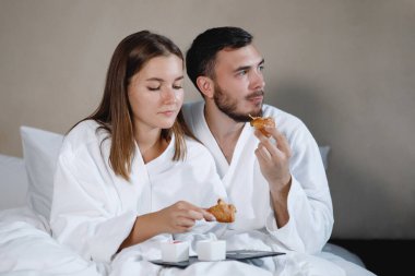 Sakallı bir adam ve beyaz önlüklü bir kadın yatakta kahvaltı yapıyorlar. Seçici odak.
