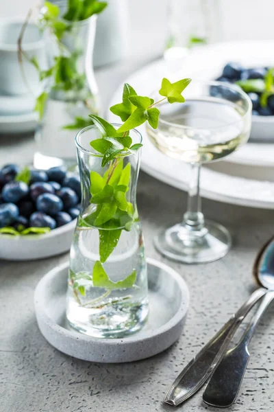 Dining room table decorated with small vase with ivy (Hedera helix). Place setting with plates and bowl with blueberries and glass of wine.