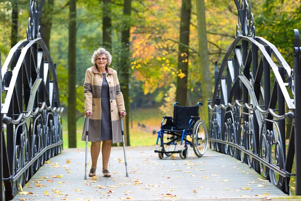 happy older woman practicing walking on crutches in autumnal park with wheelchair in the background