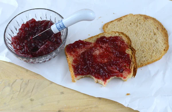 Raspberry preserves in a crystal bowl next to wheat bread spread with the preserves. All on butcher paper with a knife added. Bites have been taken of the snack.
