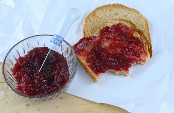 Raspberry preserves in a crystal bowl next to wheat bread spread with the preserves. All on butcher paper with a knife added. Bites have been taken of the snack.