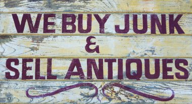 Humorous old wooden sign board saying We Buy Junk and Sell Antiques. Pale painted background with burgundy paint. clipart