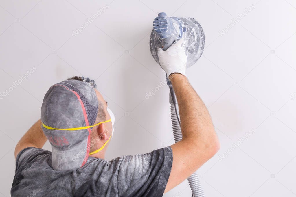 Plasterer wearing dust mask polishes a wall with sanding machine . House renovation concept.