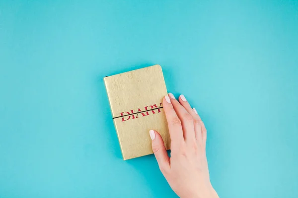 Creative flat lay of woman hands holding small golden notebook diary with copy space on turquoise paper background in minimalism style. Template for feminine blog, social media