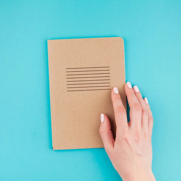 Creative flat lay of woman hands holding craft paper notebook diary with copy space on turquoise background in minimalism style. Square Template for feminine blog, social media