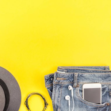 Düz mavi jeans, gri şapka ve akıllı telefon kulaklık kalın sarı Kağıt arka plan üzerinde kopya alanı ile yatıyordu. Kadın rahat kıyafet havai görünümü. Kare hipster bak üstten görünüm