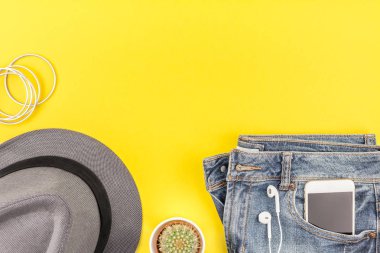 Düz mavi jeans, gri şapka, kaktüs ve akıllı telefon kulaklık kalın sarı Kağıt arka plan üzerinde kopya alanı ile yatıyordu. Kadın rahat kıyafet havai görünümü. Trendy hipster bak üstten görünüm