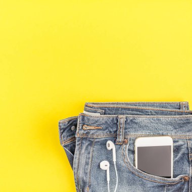 Düz mavi kot pantolon ve akıllı telefon kulaklık kalın sarı Kağıt arka plan üzerinde kopya alanı ile yatıyordu. Kadın rahat kıyafet havai görünümü. Kare hipster bak üstten görünüm