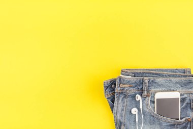 Düz mavi kot pantolon ve akıllı telefon kulaklık kalın sarı Kağıt arka plan üzerinde kopya alanı ile yatıyordu. Kadın rahat kıyafet havai görünümü. Trendy hipster bak üstten görünüm