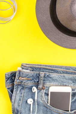Düz mavi jeans, gri şapka ve akıllı telefon kulaklık kalın sarı Kağıt arka plan üzerinde kopya alanı ile yatıyordu. Kadın rahat kıyafet havai görünümü. Trendy hipster bak üstten görünüm