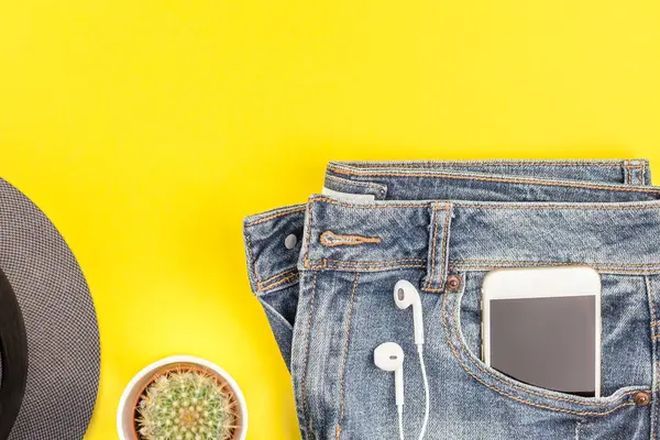 Düz mavi jeans, gri şapka, kaktüs ve akıllı telefon kulaklık kalın sarı Kağıt arka plan üzerinde kopya alanı ile yatıyordu. Kadın rahat kıyafet havai görünümü. Trendy hipster bak üstten görünüm