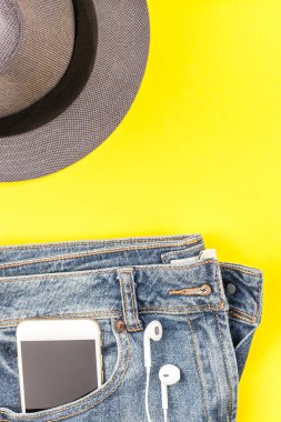 Düz mavi jeans, gri şapka ve akıllı telefon kulaklık kalın sarı Kağıt arka plan üzerinde kopya alanı ile yatıyordu. Kadın rahat kıyafet havai görünümü. Trendy hipster bak üstten görünüm