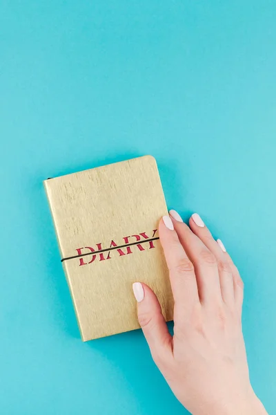 Creative flat lay of woman hands holding small golden notebook diary with copy space on turquoise paper background in minimalism style. Template for feminine blog, social media