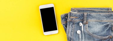 Düz mavi kot pantolon ve akıllı telefon kulaklık kalın sarı Kağıt arka plan üzerinde kopya alanı ile yatıyordu. Kadın rahat kıyafet havai görünümü. Uzun geniş afiş