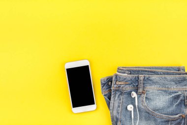 Düz mavi kot pantolon ve akıllı telefon kulaklık kalın sarı Kağıt arka plan üzerinde kopya alanı ile yatıyordu. Kadın rahat kıyafet havai görünümü. Trendy hipster bak üstten görünüm