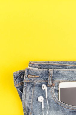 Düz mavi kot pantolon ve smartphone kalın sarı Kağıt arka plan kopya alanı ile kulaklıktan yatırın. Kadın rahat kıyafet havai görünümü. Trendy hipster bak üstten görünüm