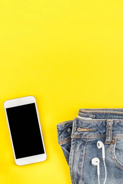 Düz mavi kot pantolon ve smartphone kalın sarı Kağıt arka plan kopya alanı ile kulaklıktan yatırın. Kadın rahat kıyafet havai görünümü. Trendy hipster bak üstten görünüm