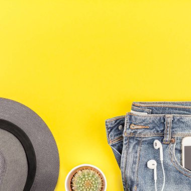 Düz mavi jeans, gri şapka, kaktüs ve akıllı telefon kulaklık kalın sarı Kağıt arka plan üzerinde kopya alanı ile yatıyordu. Kadın rahat kıyafet havai görünümü. Kare hipster bak üstten görünüm