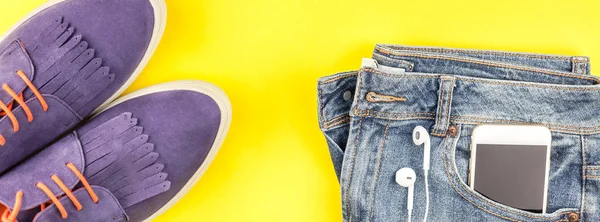 Flat lay of suede shoes, blue jeans, grey hat, cactus and smartphone headphones on bold yellow paper background with copy space. Overhead view of woman casual outfit. Long wide banner