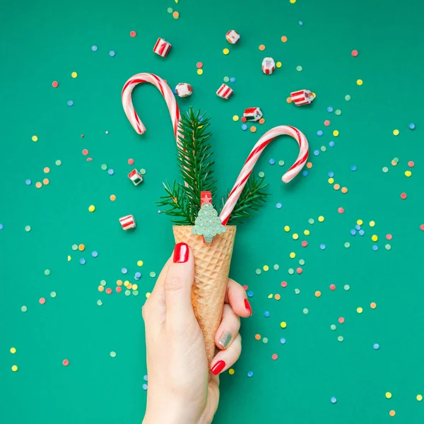 New Year Christmas Xmas holiday celebration woman hand red manicure holding waffle cone candy canes fir tree branch copy space green color paper background. Square Template greeting card 2019