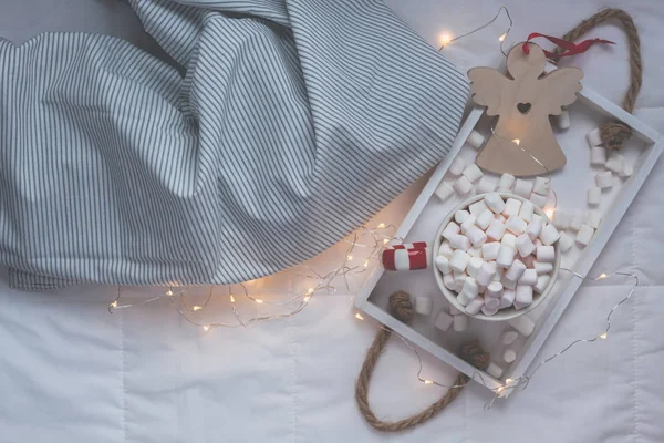 New Year or Christmas flat lay top view with Hot cacao coffee chocolate with marshmallows mug Xmas holiday celebration on wooden tray in bed with lights. Concept blog social media 2019