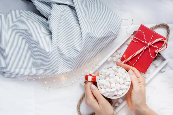 New Year or Christmas flat lay top view with Hot cacao coffee chocolate with marshmallows mug Xmas holiday celebration red present box on wooden tray in bed with lights and woman hands. Concept 2019