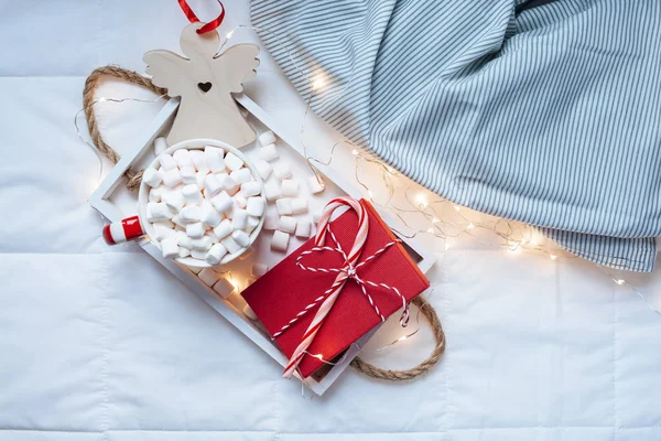 New Year or Christmas flat lay top view with Hot cacao coffee chocolate with marshmallows mug Xmas holiday celebration red present box on wooden tray in bed with lights. Concept blog social media 2019