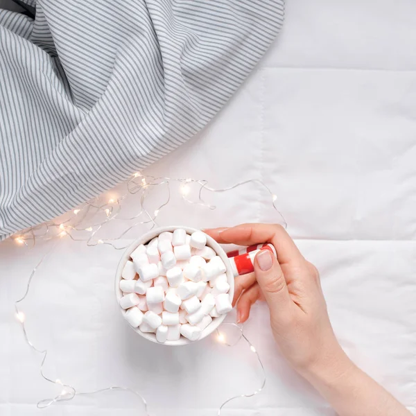 New Year or Christmas flat lay top view with Hot cacao coffee chocolate with marshmallows mug in woman hands Xmas holiday celebration in bed with lights. Square Concept blog social media 2019