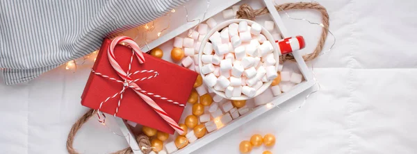 New Year or Christmas flat lay top view with Hot cacao coffee chocolate with marshmallows mug Xmas holiday celebration red present box on wooden tray in bed with lights. 2019 Long wide banner