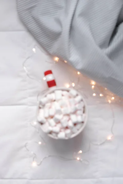 New Year or Christmas flat lay top view with Hot cacao coffee chocolate with marshmallows mug Xmas holiday celebration in bed with lights. Blurred defocused background
