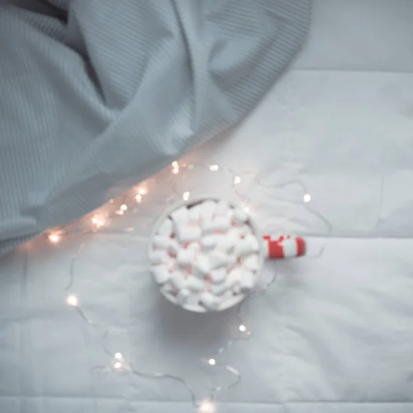 New Year or Christmas flat lay top view with Hot cacao coffee chocolate with marshmallows mug Xmas holiday celebration in bed with lights. Blurred defocused square background