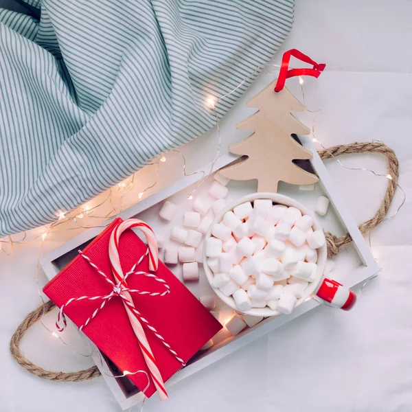 New Year or Christmas square flat lay top view with Hot cacao coffee chocolate with marshmallows mug Xmas holiday celebration red present box on wooden tray in bed with lights. blog social media 2019