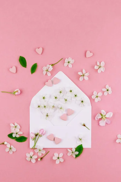 Creative flat lay concept top view of blank letter envelope, spring flowers and hearts candies on millennial pink background with copy space in minimal style, template for valentine cards